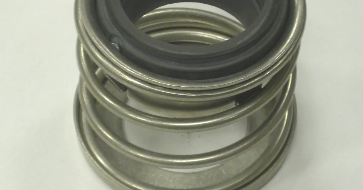 Spring-type pump mechanical seal with a grey carbon face ring inside a stainless steel retainer and coil spring, on a white surface