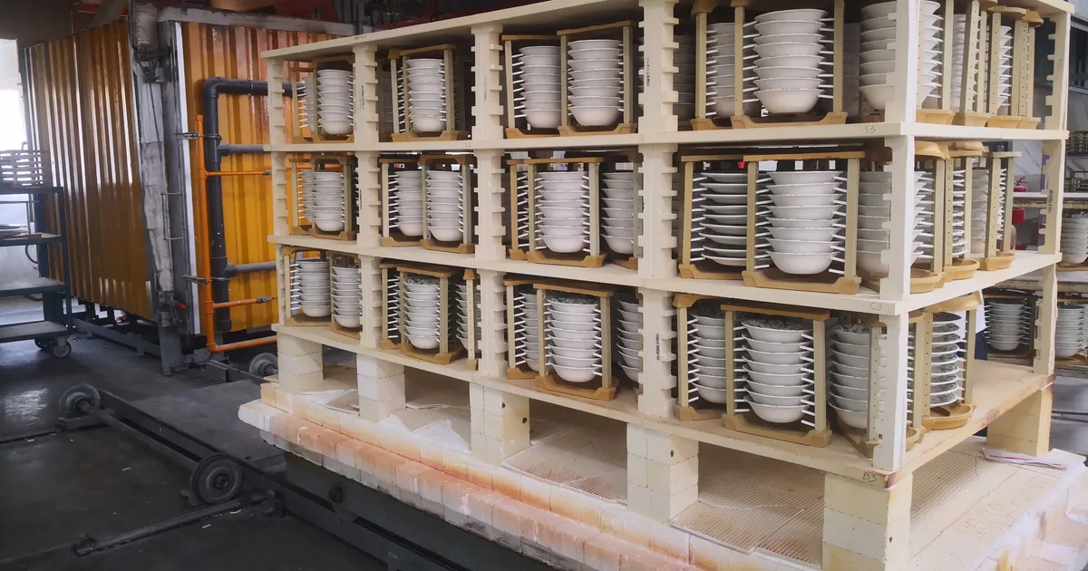 Kiln car stacked with ceramic kiln shelves, posts and setters holding rows of white bowls before a firing